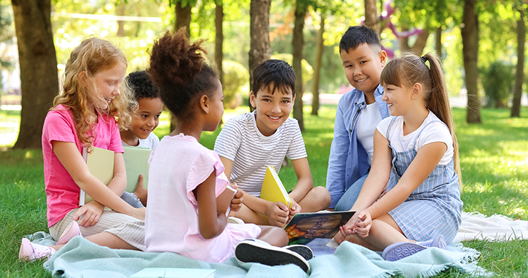 A group of school age children sitting on picnic blanket in park and reading